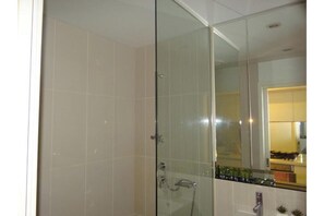 Ein durch eine Glaswand abgetrennter Duschbereich mit sichtbarer Armatur und Brause, eine geflieste Wand und ein Küchenbereich, der sich im Spiegel spiegelt.