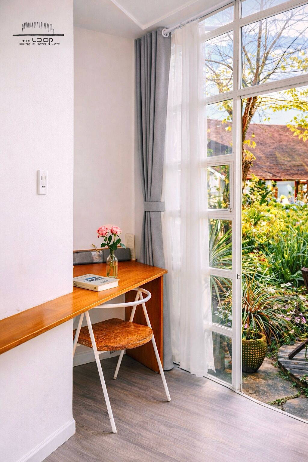 A room with a wooden desk, a chair, a vase with flowers, and a view of a garden through a large window.