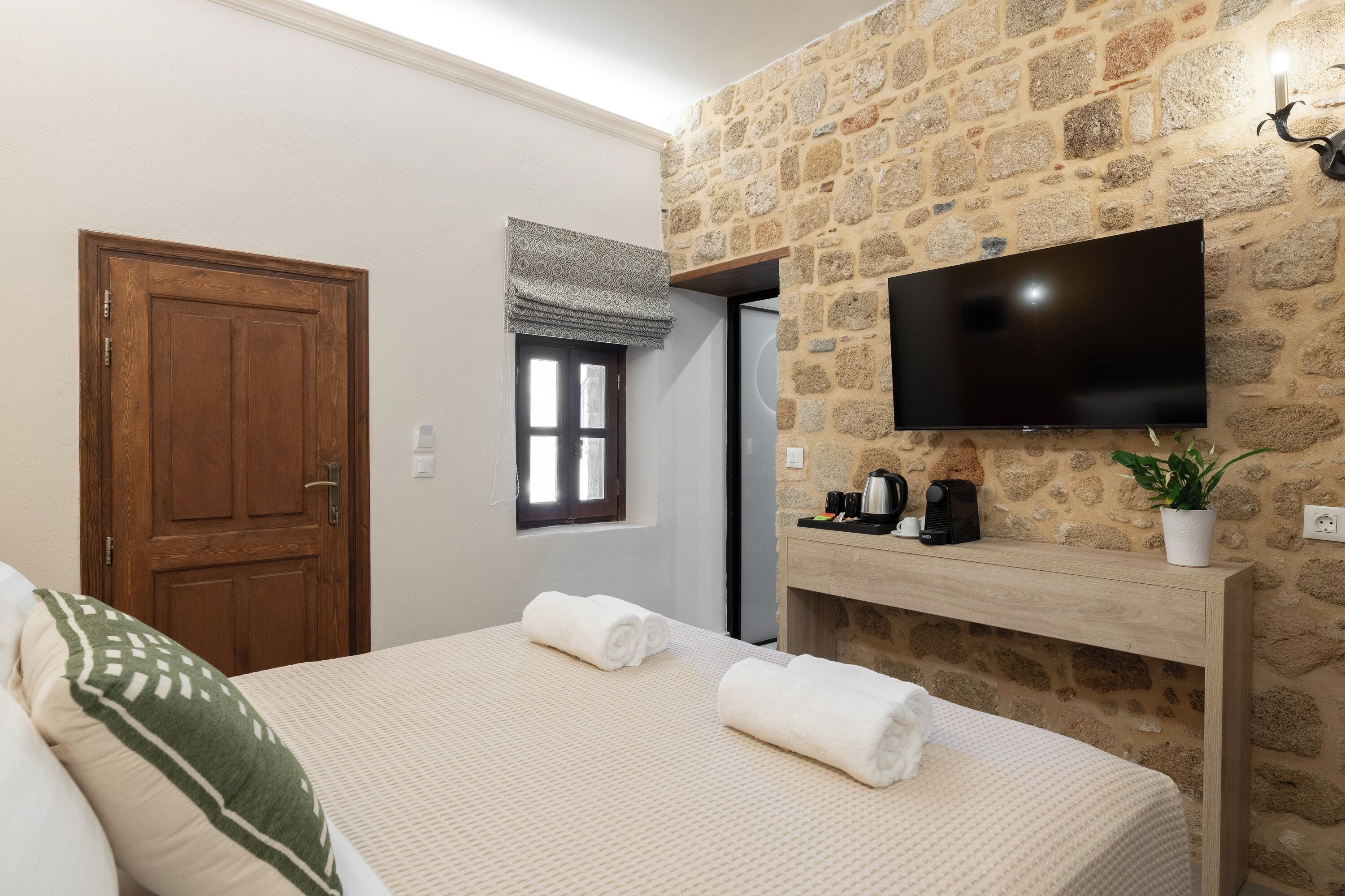 A hotel room with a stone wall, a flat-screen TV, a wooden bedside table, and a bed with rolled towels.