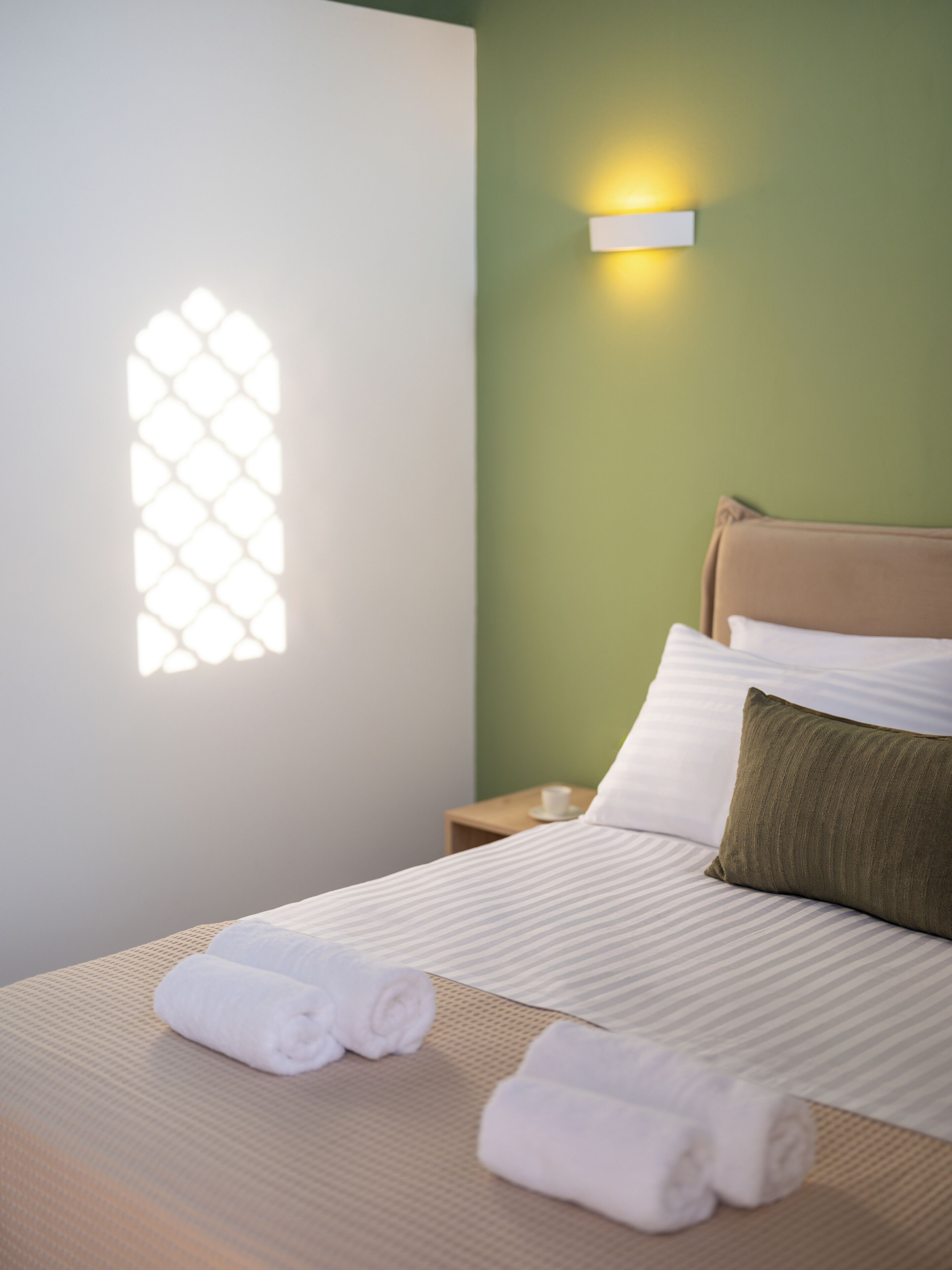 A neatly made bed with white linens and rolled towels, a green wall, and a small bedside table with a cup.