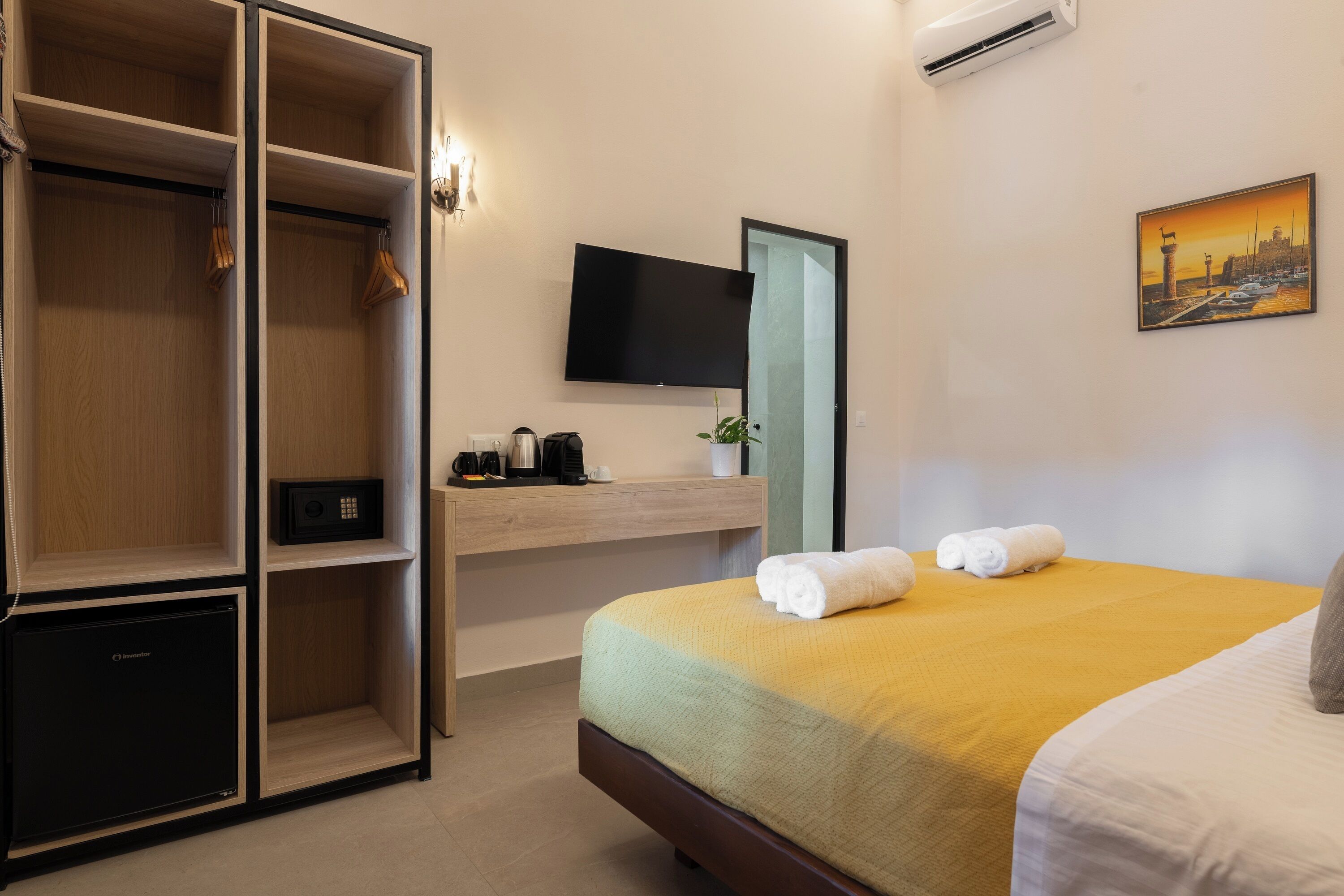 A hotel room with a bed, a wardrobe, a desk with a flat-screen TV, a small refrigerator, and a painting on the wall.