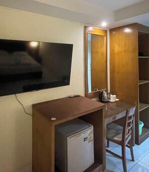 A compact room with a flat-screen TV mounted on the wall, a small refrigerator, a wooden desk, and a chair. - Bursa Hotel (Patong)