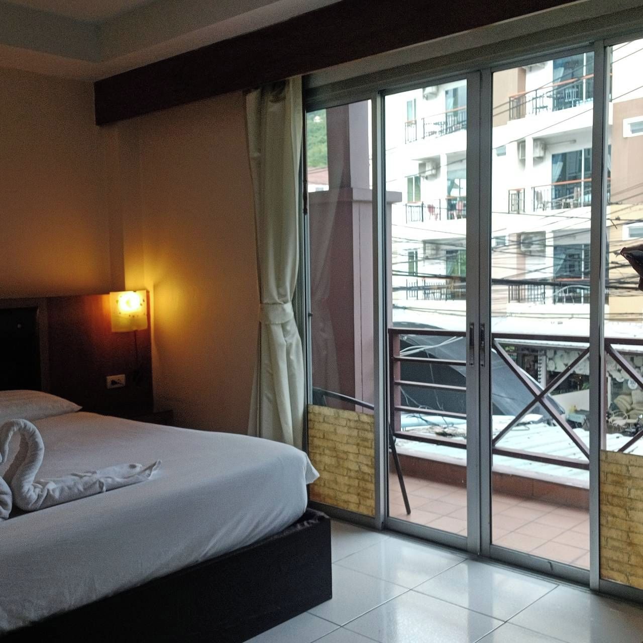 Habitación de hotel con cama, mesita de noche, lámpara y puerta corrediza de vidrio a un espacio exterior con vistas a edificios.