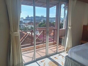 Una habitación con un amplio ventanal corredera que da a un balcón con barandilla roja, una cama con cubrecama blanco y piso de baldosas con diseño.