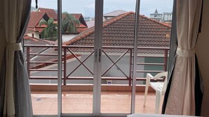 Een kamer met een groot raam, een balkon met een metalen hekwerk en uitzicht op omliggende gebouwen met rode pannendaken.