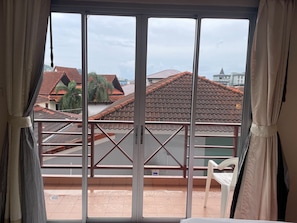 A room with a large window, a balcony with a metal railing, and a view of neighboring buildings with red-tiled roofs. - Bursa Hotel (Patong)