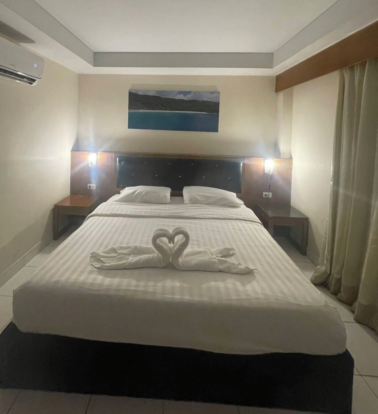 Habitación de hotel con cama, dos mesitas de noche, un buró, un cuadro en la pared y una ventana con cortinas.