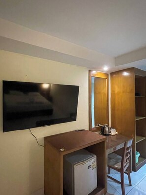 A compact room with a flat-screen TV mounted on the wall, a small refrigerator, a wooden desk, a chair, and a wooden cabinet with a mirror. - Bursa Hotel (Patong)