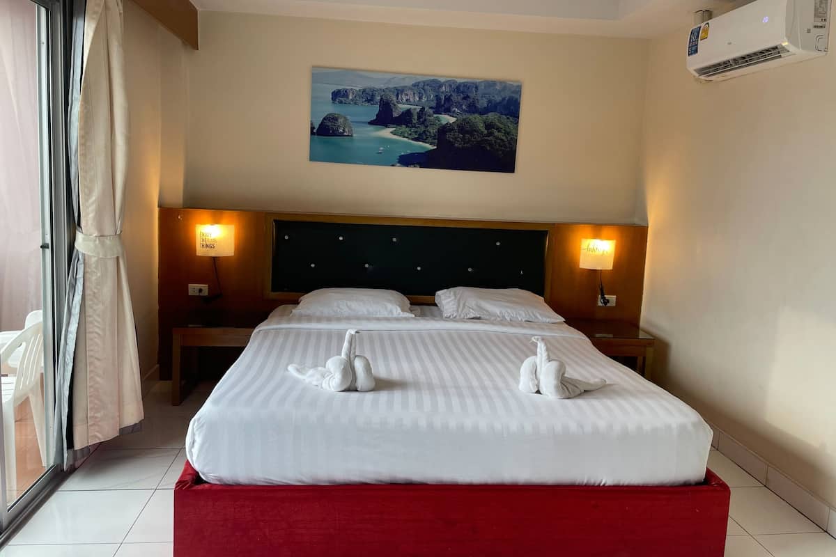 A hotel room with a large bed, two bedside lamps, a painting of a landscape, and an air conditioning unit.