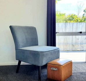 Quarto moderno com poltrona estofada cinza, cômoda de madeira, janela com vista para o verde e cortina escura.
