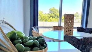Ein Zimmer mit einem runden Tisch und Korbstühlen, einer Fruchtkorb mit grünen Früchten und Blick durch große Fenster.