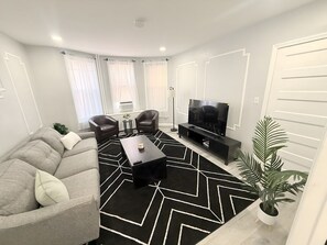 A modern living room with a sofa, armchairs, a coffee table, a TV, and a decorative plant. - New Modern JC Gem w/ On-Site Parking • Minutes to NYC, EWR & MetLife • Sleeps 6 (Jersey City)