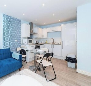 A compact kitchen with white cabinets, a microwave, and a sink. There is a blue sofa and a glass dining table with two chairs. - Free Parking Central Reading Flat Fully Equipped (Berkshire)