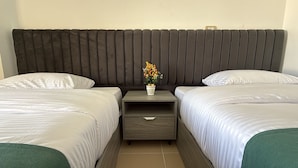 Quarto de hotel com duas camas, cabeceira escura, mesa de cabeceira com planta e chão ladrilhado.