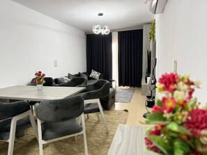 Ein modernes Wohnzimmer mit einer Sofagarnitur, einem Esstisch mit Stühlen, einer Kronleuchter und einem Fernsehmöbel mit Pflanze.