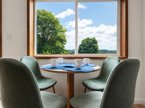 Een kamer met een ronde tafel en vier stoelen, een raam met uitzicht op bomen en de lucht.