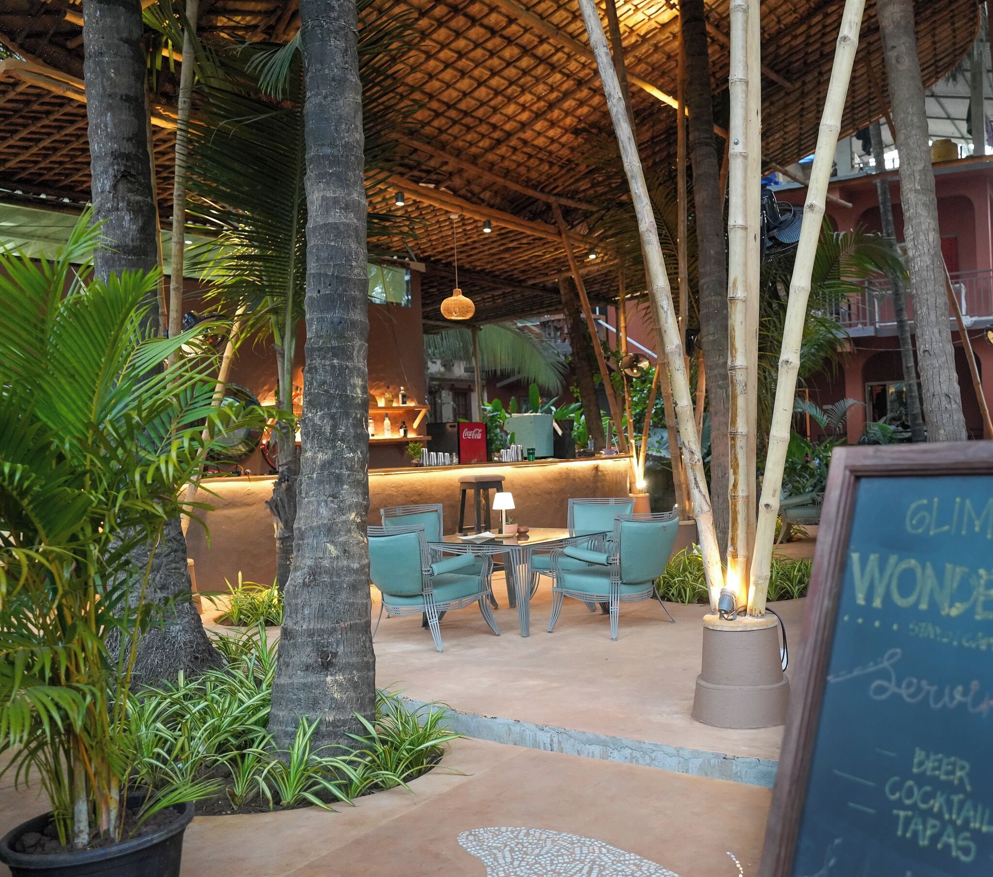 An outdoor seating area with tables and chairs, surrounded by tropical plants and a thatched roof structure.