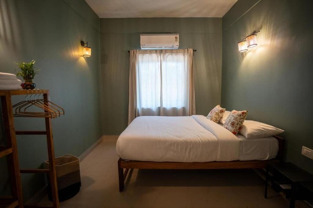 A bedroom with a bed, a wall-mounted air conditioner, a window with curtains, and a bedside table with a plant.