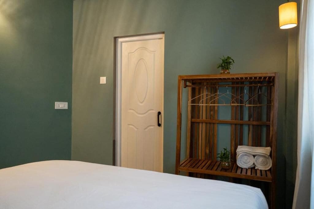 Una habitación con cama, una puerta, un estante de madera con toallas y una pequeña planta.