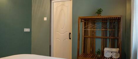 Una habitación con cama, una puerta, un estante de madera con toallas y una pequeña planta.