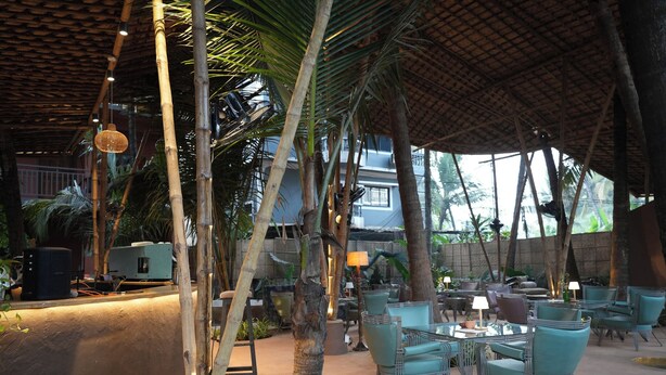 An outdoor seating area with tables and chairs, surrounded by palm trees and a thatched roof structure. - Wonderland  (Mandrem pernem)