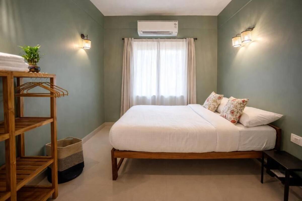 Un dormitorio con cama, estante de madera, planta colgante, aire acondicionado y ventana con cortinas.