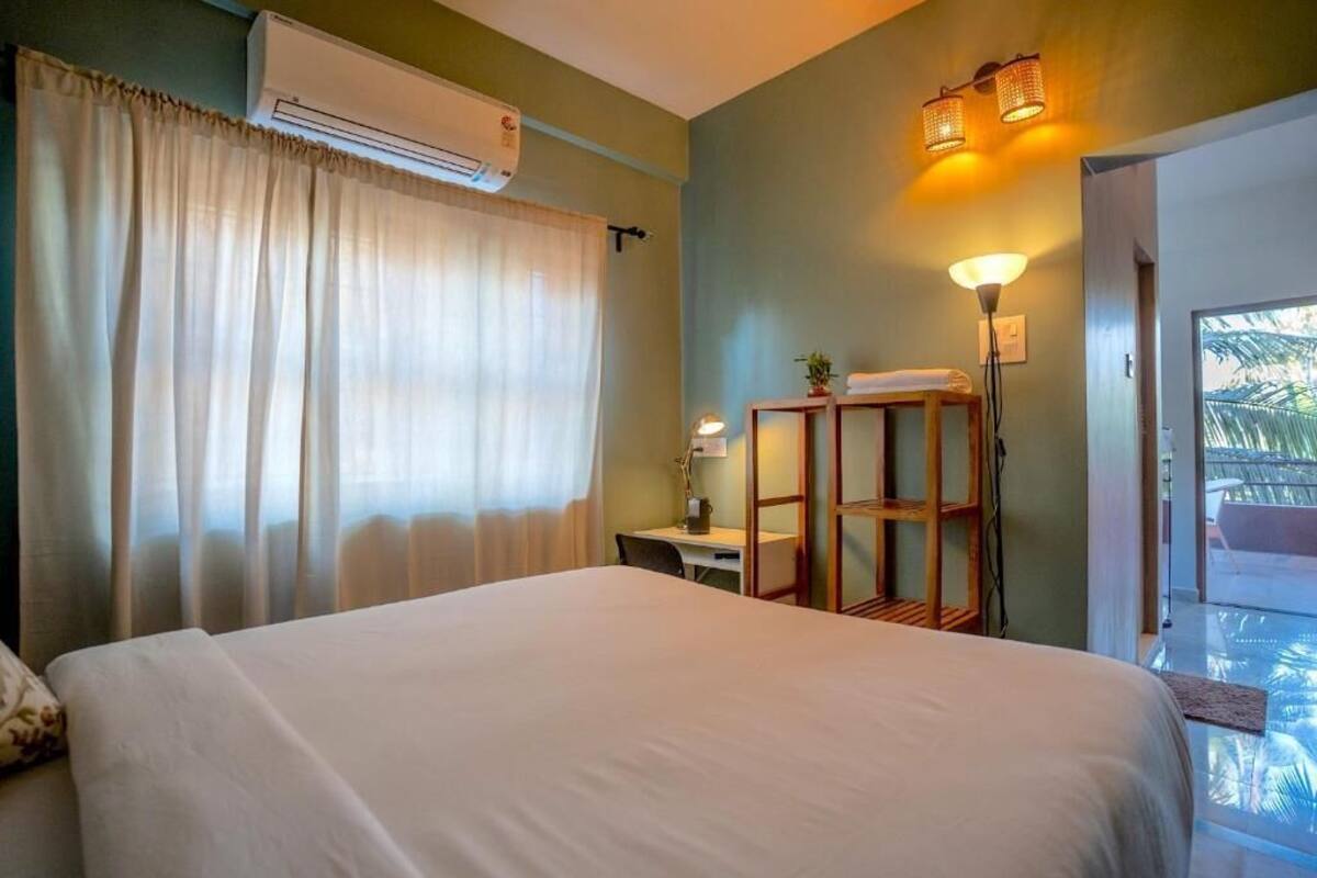 Habitación de hotel con cama, escritorio, silla, equipo de aire acondicionado, lámpara y vistas al exterior.