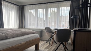 Une chambre à coucher comprenant un lit, un bureau, deux chaises et une vue sur les immeubles à travers de grandes fenêtres.