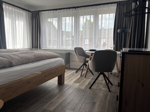 Une chambre à coucher comprenant un lit, un bureau, deux chaises et une vue sur les immeubles à travers de grandes fenêtres.