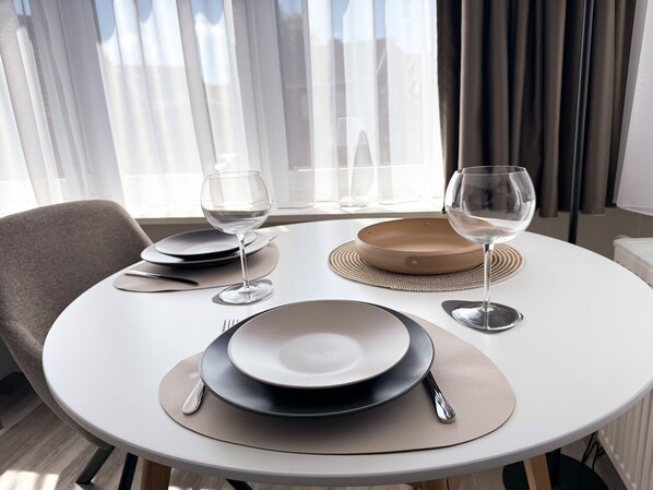 Une table de salle à manger ronde, avec assiettes, couverts et verres, placée devant une fenêtre ornée de rideaux.