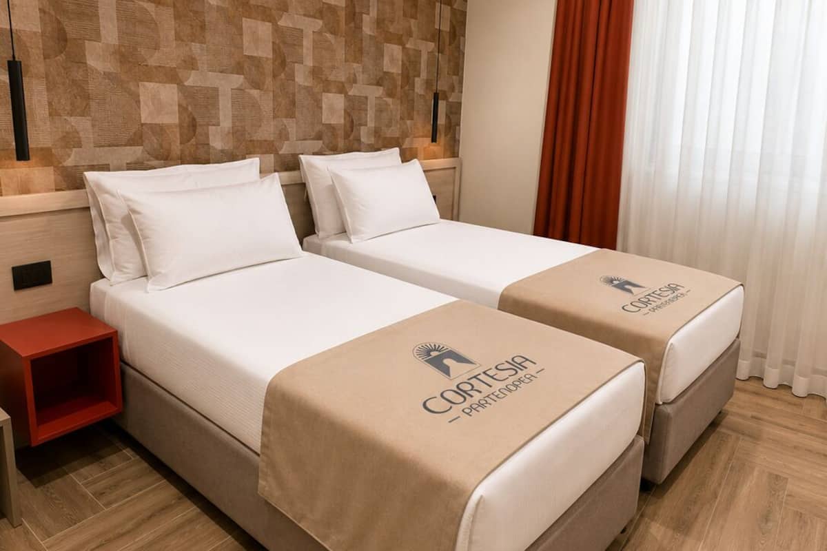 Quarto de hotel com duas camas, parede com padrão de madeira, mesa de cabeceira vermelha e janela com cortinas.