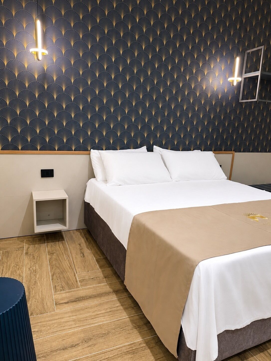 Una habitación de hotel con papel tapiz estampado, una cama con ropa de cama blanca, una mesita de noche y un suelo de madera.