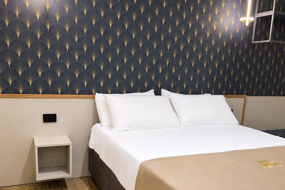 Quarto de hotel com papel de parede estampado, cama com roupa de cama branca, mesa de cabeceira e piso de madeira.
