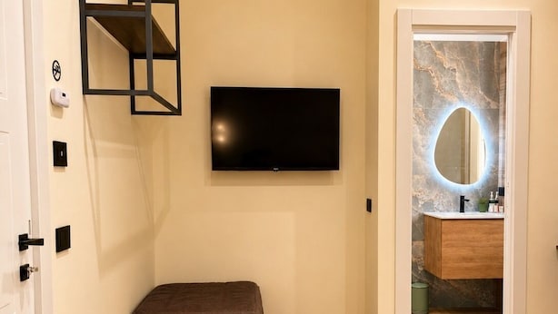 Un dormitorio moderno con una televisión de pantalla plana, un banco, una puerta y un baño con pared de mármol y un espejo circular.