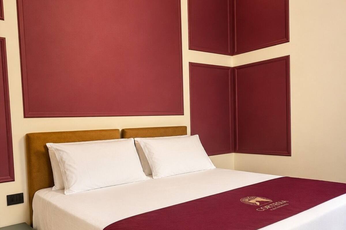 Uma cama bem arrumada com colcha branca e bordô, uma cabeceira de madeira e uma mesinha de cabeceira com abajur escuro.