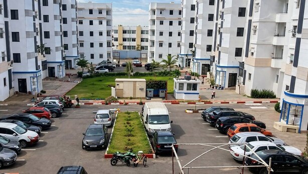Um edifício residencial de vários andares com estacionamento, uma pequena área verde e uma cabine de segurança.