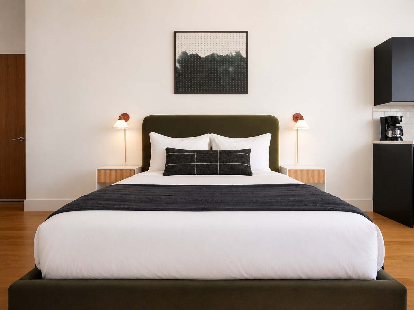 A neatly made bed with white and dark bedding, two bedside lamps, a framed picture on the wall, and a coffee machine on a cabinet.