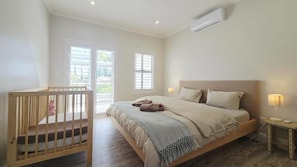 Un dormitorio con cama, cuna, mesita de noche con lámpara y ventana con persianas.