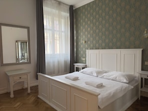 Ein Schlafzimmer mit einem großen Bett, einem Nachttisch, einem Spiegel und einem Fenster mit durchsichtigen Vorhängen.