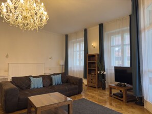 Uma sala de estar com sofá, mesa de centro, televisão, estante de livros e lustre.