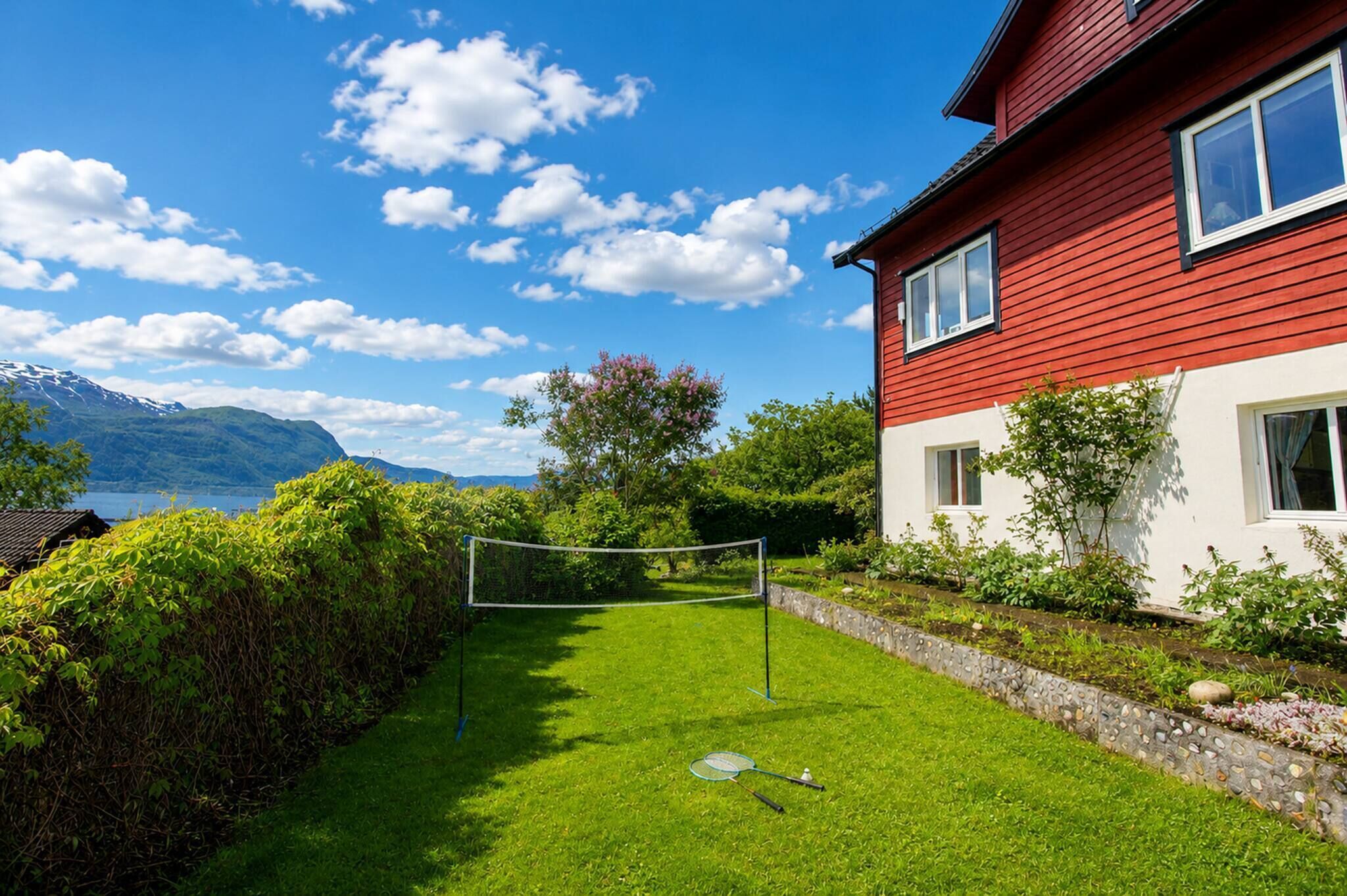 A red house with a tennis court in front, surrounded by greenery and a mountain in the background.