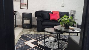 A living room with a black sofa, a red pillow, a round table with a plant, a fan, and framed pictures on the wall.