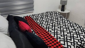 A bedroom with a bed, a window, a picture on the wall, and a small table with a lamp.
