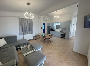 Ein modernes Wohnzimmer mit Sofa, Couchtisch und Hocker, ein Essbereich mit Stühlen sowie eine Küche mit Schränken und Geräten.