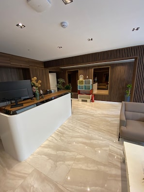Una zona de recepción moderna con suelo de mármol, paneles de madera, mostrador de recepción, un sofá y un televisor.
