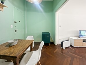 Um quarto com uma mesa de madeira, cadeiras brancas, um armário verde-água e outro armário branco com uma televisão, além de um piso de madeira.