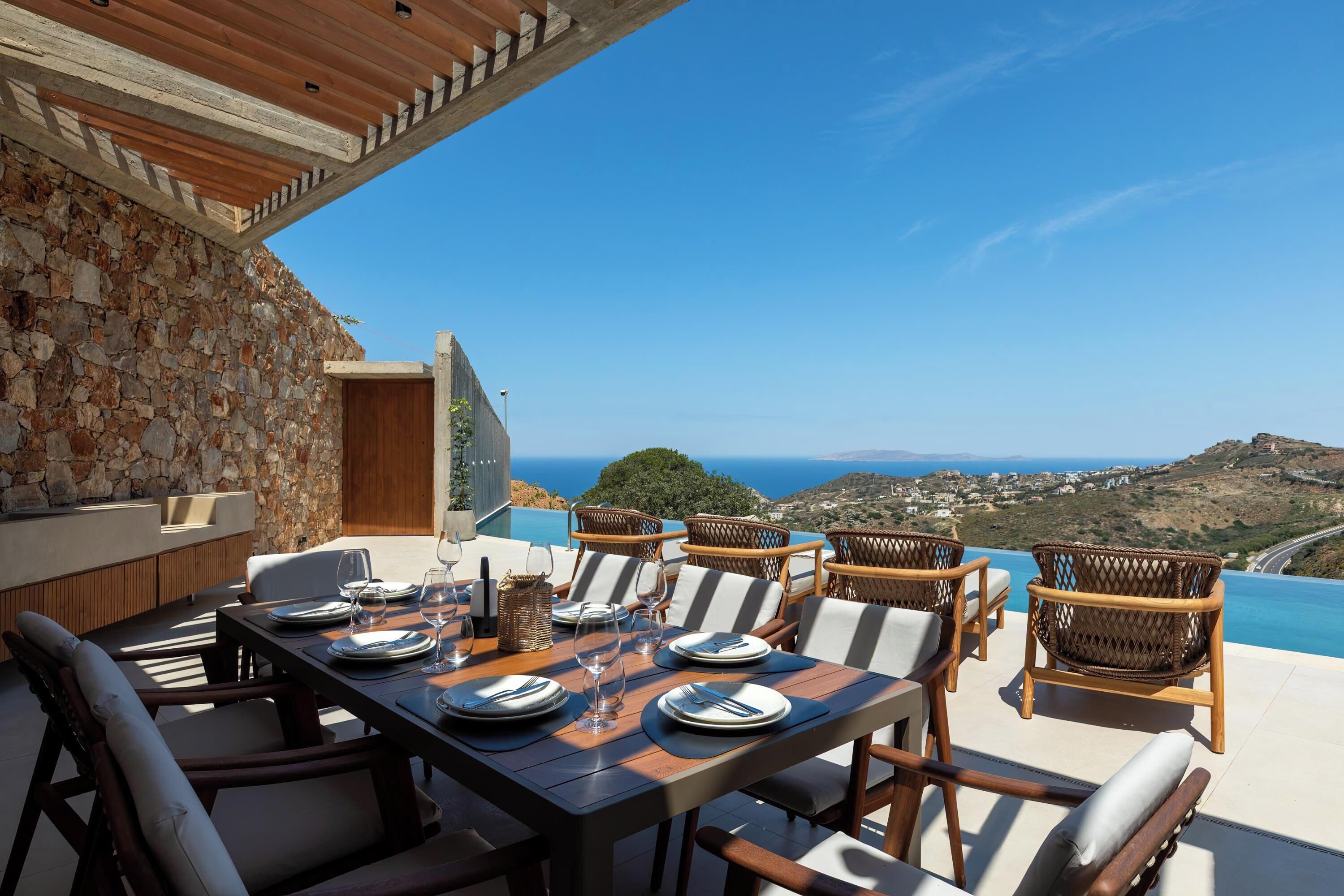 A dining area with a table set for four, wicker chairs, a stone wall, and a view of the sea.