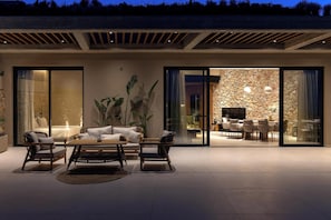An outdoor living area with a sofa, chairs, and a table, connected to a glass door leading to an interior space with a stone wall and seating. - Carved From Calm by etouri (Heraklion)