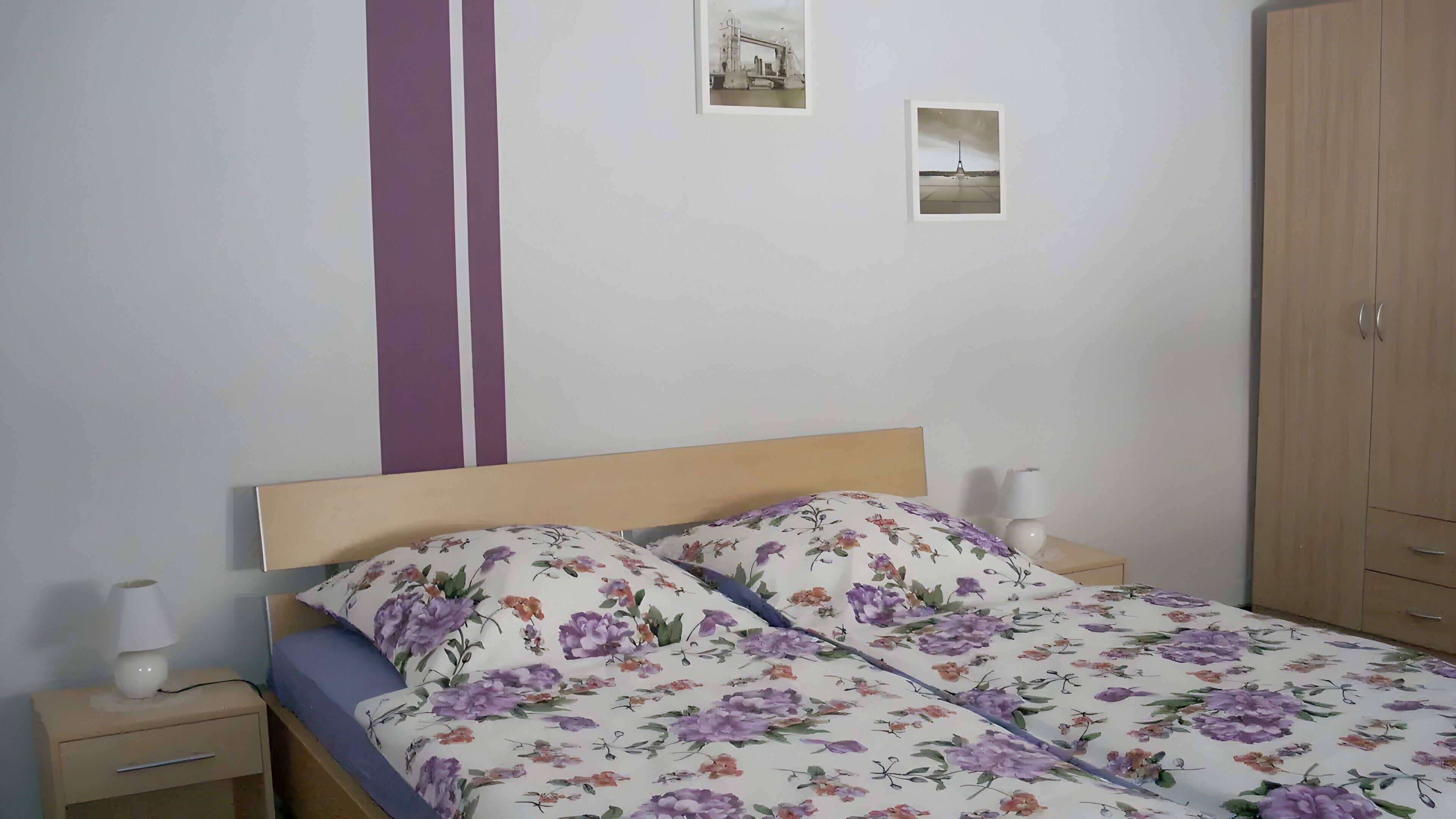 Une chambre à coucher avec une tête de lit en bois, un lit recouvert d’une literie à motifs floraux, une table de chevet avec une lampe et une armoire.