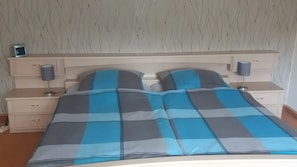 Sängyssä on raidallinen päiväpeite, sen molemmilla puolilla on yöpöydät, joissa on lamput. Seinässä on lehtikuvio, ja vasemmanpuoleisella pöydällä on pieni kello.